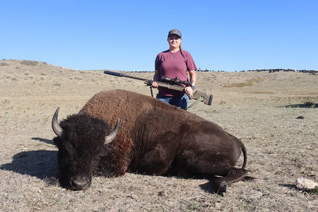 Hunter in a maroon shirt and cap holding a rifle behind her harvested bison bull on dry grassland.