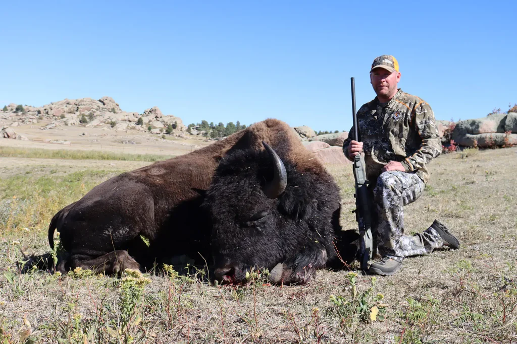 Hunter in camouflage kneeling with his rifle beside a harvested bison bull, with rocky outcrops behind.