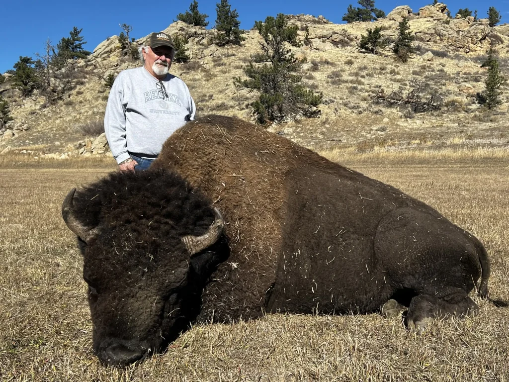 Hunter kneeling behind his harvested trophy bison bull on dry grass, with rocky hills and pines in the background.