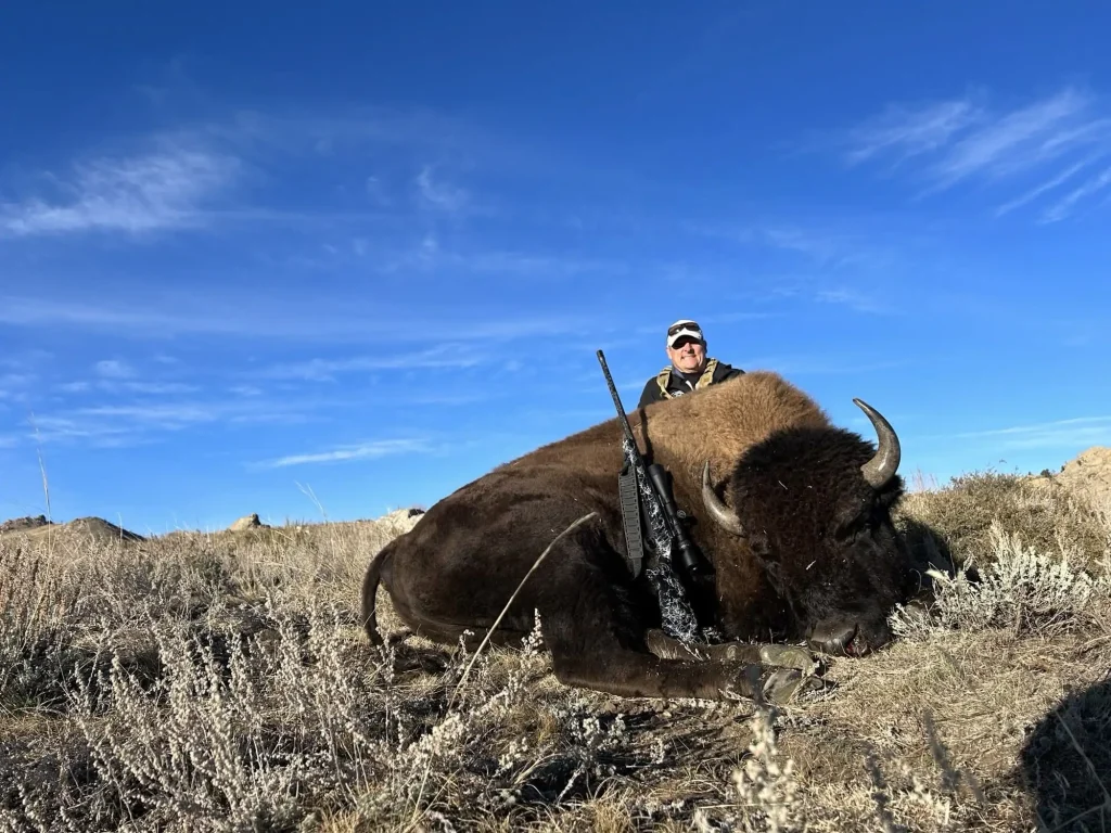 Hunter with rifle posing behind a harvested trophy bison bull on a sagebrush ridge under a clear blue sky.