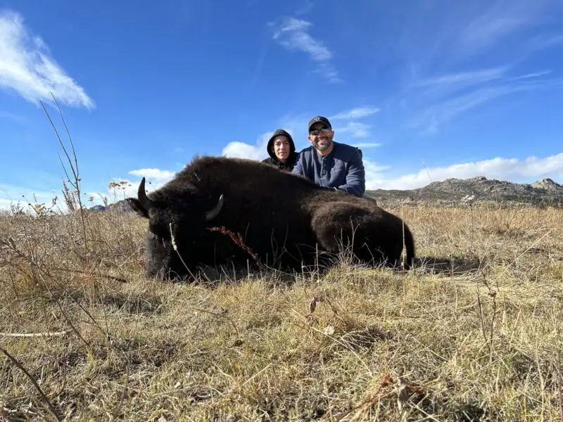 Two hunters smiling behind a harvested bison bull lying in dry grass, with scattered clouds overhead.