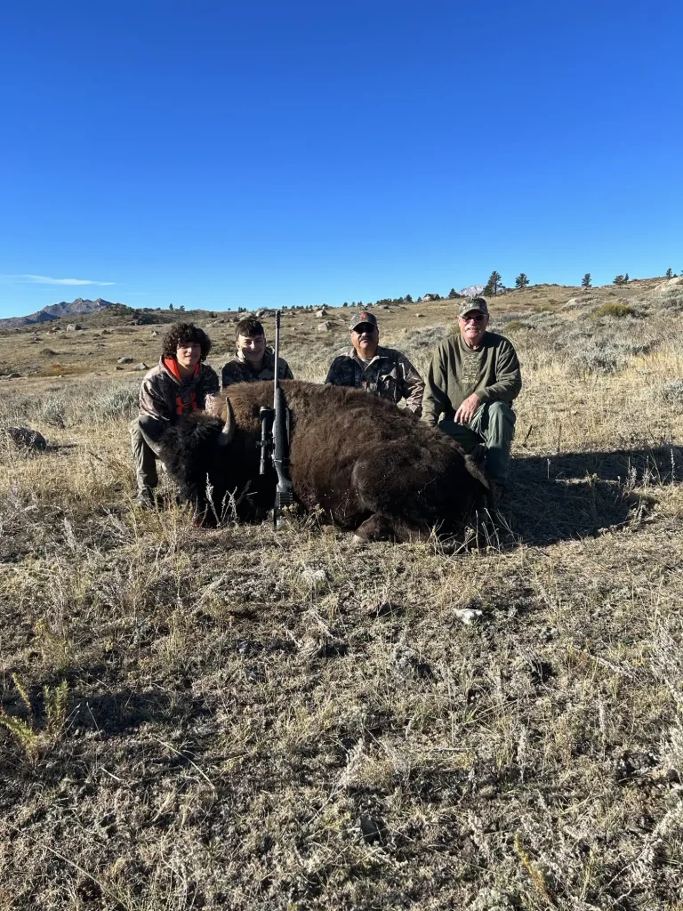 Group of four hunters gathered behind a harvested bison heifer on a grassy hillside under a clear blue sky.