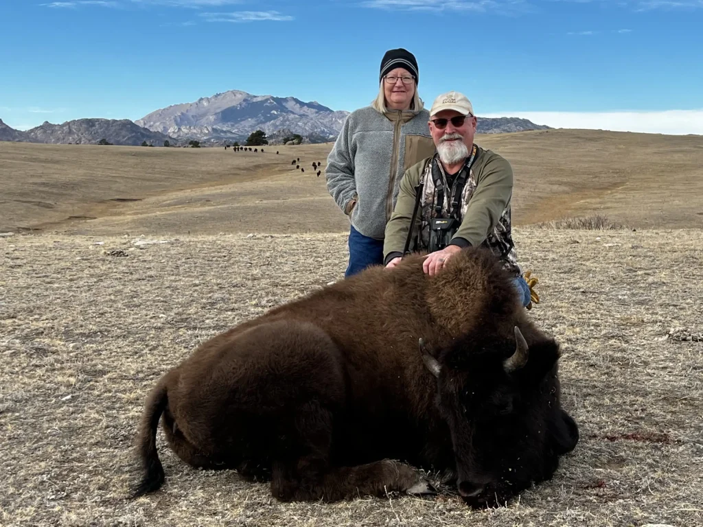 Two hunters smiling behind a harvested bison heifer, with a rugged mountain peak rising in the background.